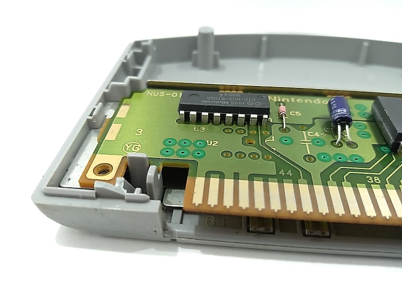 May include: Close-up of a Nintendo 64 game cartridge interior. The green circuit board features various electronic components, including chips, resistors, and capacitors. The cartridge is partially encased in a gray plastic shell.