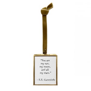 May include: A gold-toned metal frame with a white card inside. The card features a quote by E.E. Cummings that reads, "You are my sun, my moon, and all my stars." The frame is suspended from a gold ribbon.