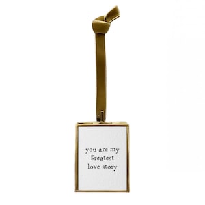 May include: A gold-toned metal frame with a white background and the text "you are my greatest love story" hanging from a gold ribbon.