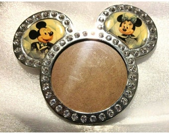 May include: Silver-toned Mickey Mouse picture frame with rhinestones. The frame has two smaller circles with images of Mickey and Minnie Mouse in formal attire. The larger circle in the center is empty for a photo.