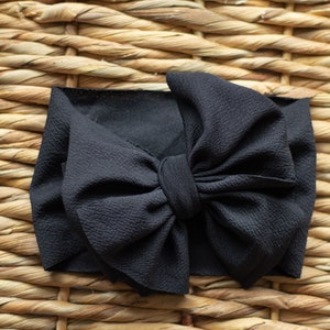 May include: A black fabric headband with a large bow. The headband is made of a textured material and is knotted in the centre to form the bow. The bow is the focal point of the headband.