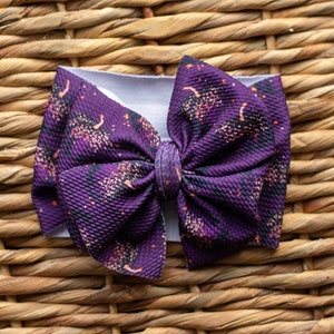 May include: A purple headband with a large bow featuring a black and orange print of trees and stars. The bow is made of a soft, textured fabric.