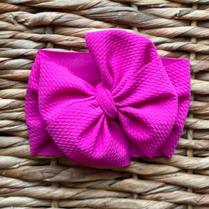 May include: A bright pink fabric headband with a large, knotted bow. The headband is made of a soft, textured fabric.