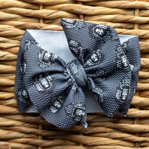 May include: A grey bow with a black and white print of a woman's face. The bow is made of a soft, textured fabric.
