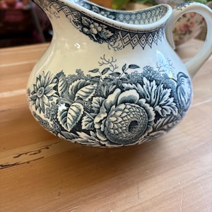 Può includere: Una brocca in ceramica con una base color crema e un motivo a trasferimento blu scuro. Il design presenta intricati motivi floreali, inclusi grandi fiori e foglie, con un bordo geometrico lungo l'apertura e il manico.