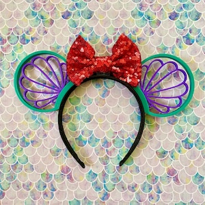 May include: A black headband with attached mermaid-themed ears and a red sequined bow. The ears are green with purple shell designs. The bow is a vibrant red, adding a pop of color to the accessory.