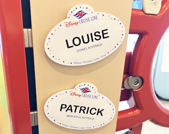Könnte beinhalten: Zwei ovale, weiße Namensschilder mit dem Disney Cruise Line-Logo und den Namen "LOUISE" und "PATRICK" in Schwarz. Die Schilder enthalten auch die Orte "SYDNEY, AUSTRALIA" und "NEWCASTLE, AUSTRALIA". Die Schilder befinden sich auf einer hellbeige Oberfläche.