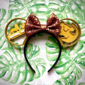 May include: A black headband with gold-coloured ears and a brown sequined bow. One ear has a lion silhouette with the text "Hakuna Matata", and the other ear has a lion cub design. The background is a green and white leaf pattern.
