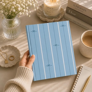 Op de afbeelding: Een lichtblauw notitieboek met harde kaft, voorzien van een patroon van verticale witte strepen en dunne donkerblauwe lijnen, geaccentueerd met kleine, gestileerde strikjes. Het notitieboek heeft een strak, gestructureerd ontwerp.