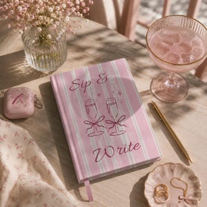 Op de afbeelding: Een roze en wit gestreept notitieboek met harde kaft, voorzien van de tekst Sip & Write en een illustratie van twee champagneglazen met strikjes. Het notitieboek heeft een roze leeslint en ligt naast een gouden pen en een roze drankje.