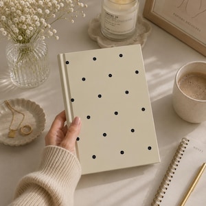 Op de afbeelding: Een crèmekleurig notitieboek met harde kaft en een herhalend patroon van kleine, zwarte stippen. Het notitieboek is rechthoekig met een gladde afwerking en een stevige rug, uitgevoerd in een neutrale kleur voor dagelijks gebruik.