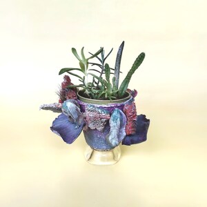 May include: A decorative planter featuring a selection of succulents and cacti. The pot is adorned with purple, blue, and red tones, set on a clear glass base. The arrangement showcases various green succulents and cacti, forming a distinctive display.