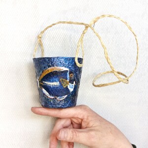 May include: A small blue hanging planter with a painted fish design. The planter is made of paper mache and has a natural fiber rope handle.