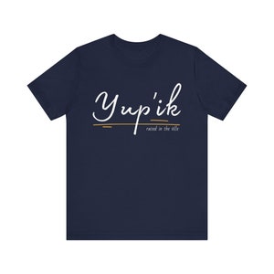 May include: A navy blue t-shirt with the text "Yup'ik" in white cursive writing and the phrase "raised in the ville" in smaller white text below.