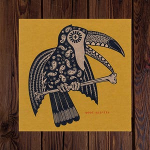 May include: A square print featuring a stylized toucan illustration in black and white paisley patterns on a mustard yellow background. The toucan holds a bone in its beak, with the words "good spirits" in red.