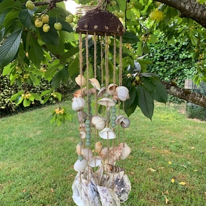Peut inclure: Un carillon de coquillages artisanal suspendu à une branche d'arbre. Le carillon est composé d'une coque de noix de coco marron, de ficelle naturelle, de divers coquillages et de perles vert clair. L'arrière-plan montre de l'herbe verte et du feuillage.
