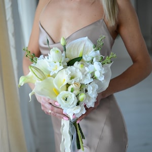 Puede incluir: Un ramo de novia con calas blancas, rosas y otras flores blancas con detalles verdes. El ramo está atado con una cinta blanca. La persona lleva un vestido de satén color champán.