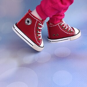 May include: Red high-top trainers with white laces and soles. The trainers have a star logo on the side and are worn with pink trousers. The shoes are approximately 5 cm in height.