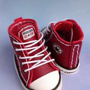 Könnte beinhalten: Ein Paar Miniatur-High-Top-Sneaker in Rot von Converse All Star. Die Schuhe haben weiße Schnürsenkel, eine weiße Gummikappe und das Converse All Star-Logo auf der Zunge. Die Sohlen sind weiß mit einem schwarzen Streifen.