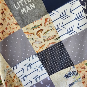 May include: A patchwork quilt with various fabric patterns. The quilt features squares with text that reads "LITTLE MAN", arrows, fish, deer, and a map. The color palette includes navy, gray, beige, and white.