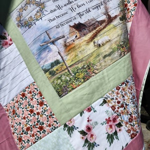 May include: A patchwork quilt featuring a central panel with a religious scene and text. The quilt incorporates various floral and patterned fabrics in shades of pink, green, and white. The text reads "I still believe in Amazing Grace".