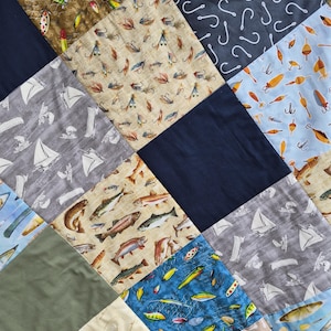 May include: A patchwork quilt with a variety of fishing-themed fabric squares. The squares feature images of fishing lures, hooks, fish, and sailboats in shades of blue, green, brown, and beige. The quilt is a mix of patterns and solid colors.