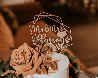 May include: A white cake with gold accents and a brown rose on top. The cake is on a wooden cake stand and has a wooden topper that says "Mr. & Mrs. Parsons" with a heart.