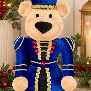 May include: A plush teddy bear dressed as a nutcracker, featuring a blue velvet uniform with gold braided trim, red gemstone accents, and a tall blue hat. The bear has tan fur, a black nose, and black eyes, with a dark green plaid fabric visible.