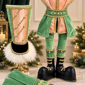May include: A decorative nutcracker costume set featuring a tall green and pink velvet hat with gold trim and white faux fur, a matching green and pink velvet tunic with gold fringe, and black boots with gold accents and black gloves.