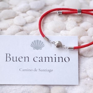 May include: A red cord bracelet with silver-tone metal accents and a seashell charm. The bracelet is displayed on a white card with the words "Buen camino" and "Camino de Santiago" printed in black.