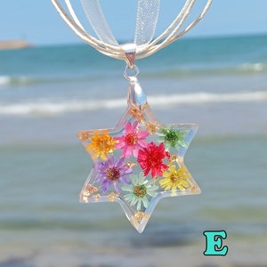 Puede incluir: Un collar de cadena de plata con un colgante de resina transparente en forma de estrella de seis puntas. El colgante contiene flores prensadas en tonos de rosa, amarillo, verde y morado.