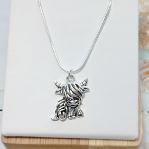May include: A silver-toned necklace featuring a pendant shaped like a Highland cow with long, shaggy hair, curved horns, and a detailed face. The pendant hangs from a thin, smooth silver-toned chain, creating a simple and clear design.