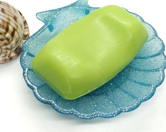 Puede incluir: Una pastilla de jabón verde claro descansando en un jabonera azul en forma de concha marina. La jabonera tiene un acabado brillante.