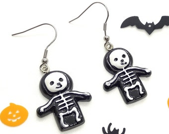 Puede incluir: Un par de pendientes colgantes con figuras de esqueleto en blanco y negro con cabezas encapuchadas y detalles visibles de la caja torácica. Los pendientes cuelgan de ganchos de metal plateado y se acompañan de pequeñas formas de calabaza naranja.