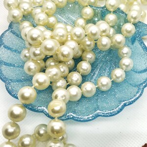Può includere: Una collana di perle finte bianche in un piatto a forma di conchiglia blu con glitter.