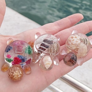 Può includere: Tre piccole figurine di tartaruga in resina trasparente con diversi design. La prima tartaruga ha una varietà di fiori e foglie colorati all'interno. La seconda tartaruga ha una varietà di piccole conchiglie all'interno. La terza tartaruga ha una varietà di piccole conchiglie e una pietra marrone all'interno.