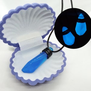 Puede incluir: Un collar con un colgante de cristal azul que brilla en la oscuridad, presentado en una caja de terciopelo morado en forma de concha. El collar tiene un cordón negro y el cristal tiene forma de lágrima.