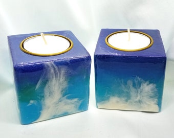 Puede incluir: Dos portavelas cuadrados de color azul verdoso, blanco y morado con bordes dorados. Cada portavelas tiene una vela de té blanca.