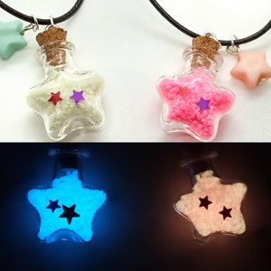 May include: Two star-shaped glass bottles filled with glow-in-the-dark sand. The bottles are on necklaces and have cork stoppers. One bottle is filled with white sand and the other is filled with pink sand. The bottles are glowing blue and pink in the dark.