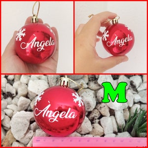May include: Red Christmas ornament with the name "Angela" in white script, accented with white snowflake designs. The ornament has a gold-colored string for hanging. The letter "M" is in green.
