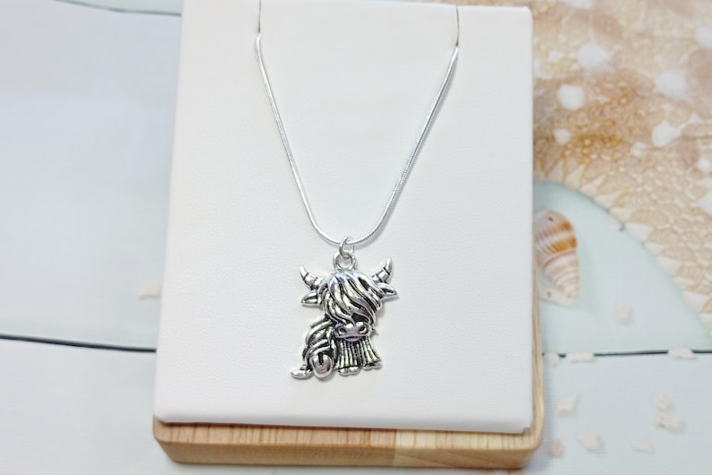 May include: A silver-toned necklace featuring a pendant shaped like a Highland cow with long, shaggy hair, curved horns, and a detailed face. The pendant hangs from a thin, smooth silver-toned chain, creating a simple and clear design.