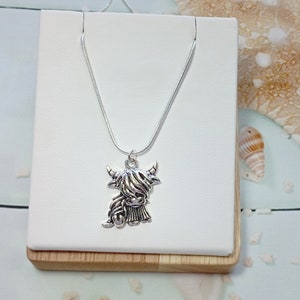 May include: A silver-toned metal necklace featuring a pendant shaped like a Highland cow with long, shaggy hair and curved horns. The pendant hangs from a thin, smooth silver-toned snake chain, creating a simple and cohesive metallic accessory.