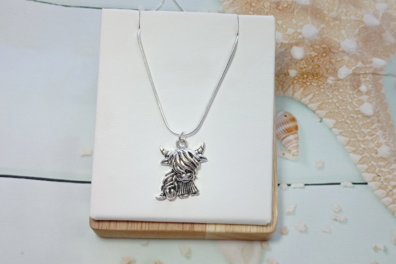 May include: A silver-toned metal necklace featuring a pendant shaped like a Highland cow with long, shaggy hair and curved horns. The pendant hangs from a thin, smooth silver-toned snake chain, creating a simple and cohesive metallic accessory.