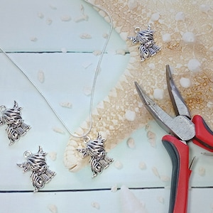 May include: A silver-toned necklace featuring a Highland cow charm, accompanied by three loose matching charms and a pair of needle-nose pliers with red and black handles. The charms depict a shaggy-haired cow with prominent horns.