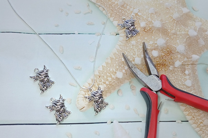 May include: A silver-toned necklace featuring a Highland cow charm, accompanied by three loose matching charms and a pair of needle-nose pliers with red and black handles. The charms depict a shaggy-haired cow with prominent horns.