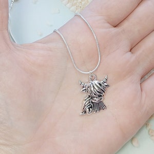 May include: A silver-toned necklace featuring a pendant shaped like a Highland cow with long, shaggy hair and curved horns. The pendant hangs from a thin, flexible silver-toned snake chain, showcasing a metallic finish and detailed texture.
