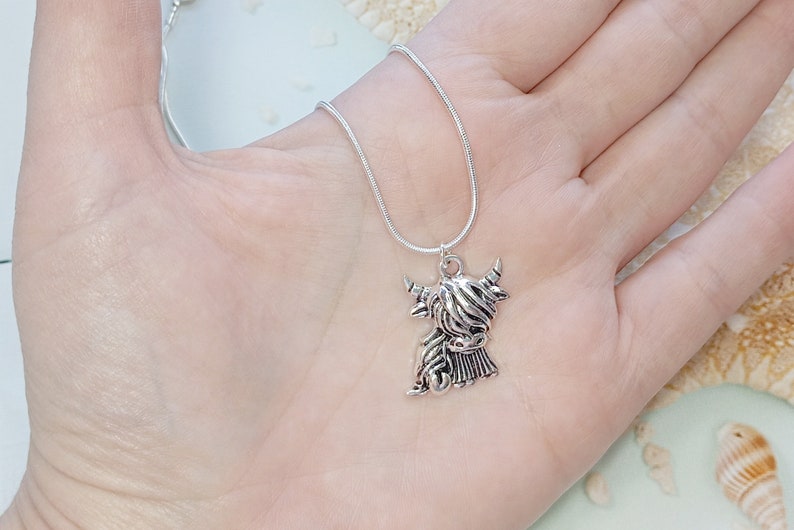May include: A silver-toned necklace featuring a pendant shaped like a Highland cow with long, shaggy hair and curved horns. The pendant hangs from a thin, flexible silver-toned snake chain, showcasing a metallic finish and detailed texture.