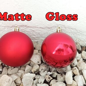 May include: Two red Christmas ornaments, one matte and one glossy, are displayed against a white wall and a bed of small white rocks. The word "Matte" is above the matte ornament, and "Gloss" is above the glossy one.