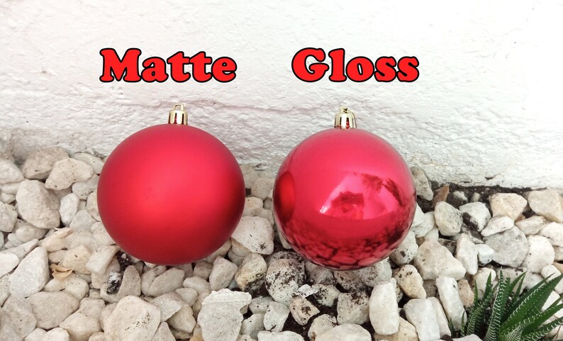May include: Two red Christmas ornaments, one matte and one glossy, are displayed against a white wall and a bed of small white rocks. The word "Matte" is above the matte ornament, and "Gloss" is above the glossy one.