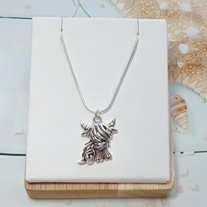 May include: A silver-toned necklace featuring a pendant shaped like a Highland cow with long, shaggy hair and curved horns. The pendant hangs from a thin, smooth silver-toned chain, creating a detailed and metallic accessory for everyday wear.